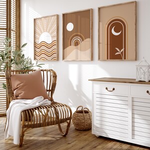 Op de afbeelding: Drie abstracte kunstprints in bruine, beige en witte tinten met geometrische vormen en ontwerpen. De prints zijn ingelijst in natuurlijk hout en hangen aan een witte muur. Een rieten stoel met een bruin kussen staat voor de prints.