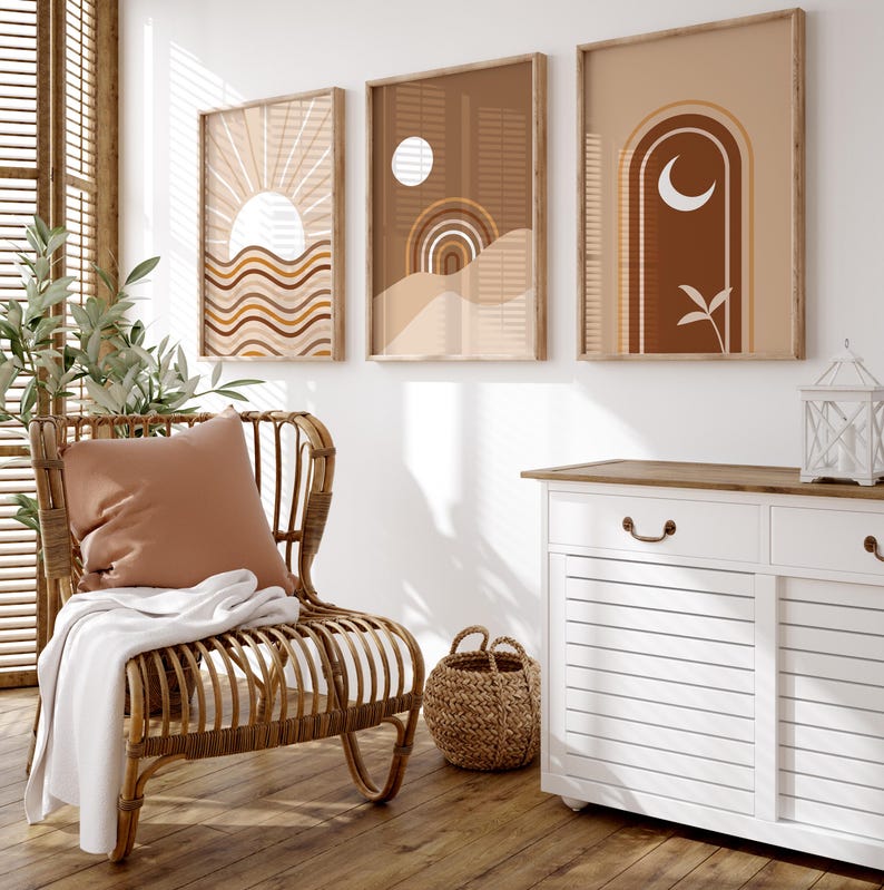 Op de afbeelding: Drie abstracte kunstprints in bruine, beige en witte tinten met geometrische vormen en ontwerpen. De prints zijn ingelijst in natuurlijk hout en hangen aan een witte muur. Een rieten stoel met een bruin kussen staat voor de prints.