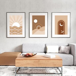 Op de afbeelding: Drie ingelijste abstracte kunstprints met een minimalistisch ontwerp met geometrische vormen en kleuren in tinten bruin, beige en wit. De prints tonen een zon, een maan en een boog.