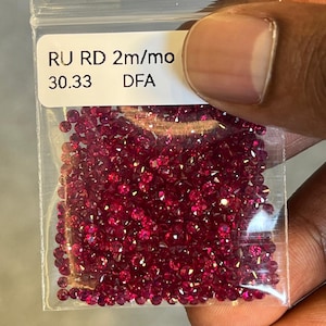 May include: A transparent plastic bag filled with many small, round, deep red gemstones. The bag has a white label with the text "RU RD 2m/mo" and "30.33 DFA". The gemstones are densely packed, creating a sparkling effect.