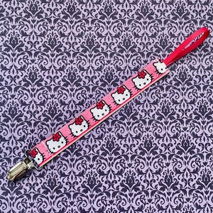May include: A pink and white pacifier clip with a repeating pattern of cartoon characters. The clip has a silver metal clasp.
