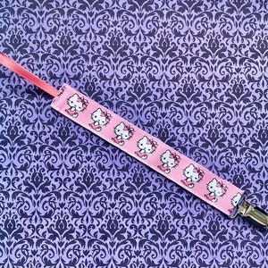 May include: Pink and white pacifier clip with a repeating pattern of Hello Kitty characters. The clip is made of fabric and has a metal clasp.