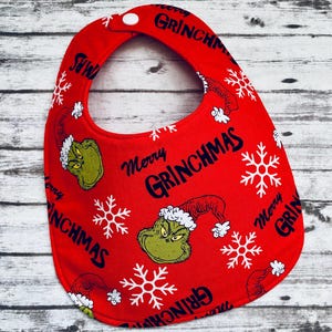 May include: Red baby bib with a white snowflake and green Grinch pattern. The bib has the text "Merry Grinchmas" repeated on it.