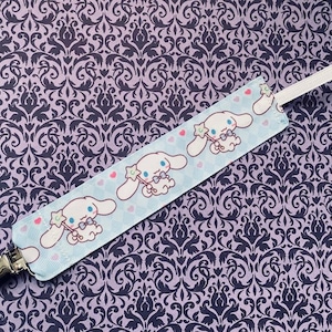 May include: A blue and white fabric baby pacifier clip with a white ribbon and a silver clip. The fabric features a repeating pattern of a cartoon dog with a pink bow and a star.