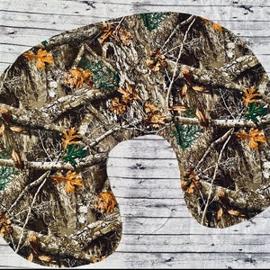 Puede incluir: Una almohada para el cuello con un patrón de camuflaje en tonos marrón, verde y beige, con ramas y hojas. La almohada está diseñada para adaptarse alrededor del cuello.