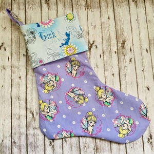 May include: Purple Christmas stocking with a white polka dot pattern and a Tinkerbell design. The stocking has a blue and white cuff with the name "Tink" embroidered on it.