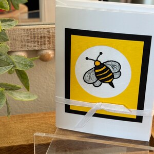 May include: A white card with a black border and a yellow square inside. The yellow square has a white circle with a black and yellow bee inside. The bee has black wings and a white body with black stripes. A white ribbon is tied around the card.