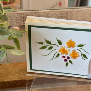 May include: A stack of blank greeting cards with a green border and a white ribbon. The cards feature an embroidered design of yellow flowers and green leaves.