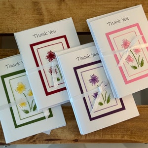 May include: Four sets of thank you cards with different colored borders and embroidered flowers. The cards are tied with white ribbon and have the text "Thank You" printed on the front.