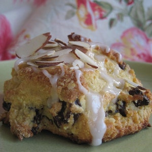 May include: A close-up of a sweet, glazed scone topped with sliced almonds. The scone is light brown and has a slightly crispy texture. It is covered in a white glaze and has a few dark raisins visible.