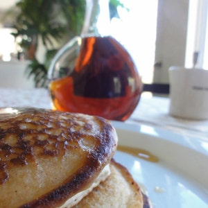 May include: A stack of three golden brown pancakes on a white plate. The pancakes are slightly burnt on the edges. A clear glass bottle of syrup is in the background.