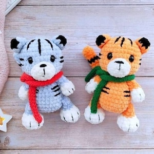 May include: Two crocheted tiger plushies. One is light blue with a red scarf, the other is orange with a green scarf. Both have black stripes, black ears, and white paws. They are on a light wood surface.