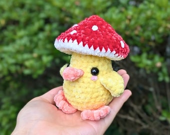 May include: A crocheted yellow chick wearing a red and white mushroom hat. The chick has a small pink beak and black eyes.