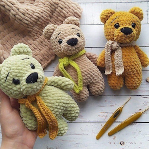 May include: Three crocheted teddy bears with different coloured fur. The bears are wearing scarves and are sitting on a white wooden surface.