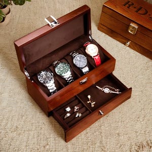 May include: A wooden watch box with a hinged lid and a drawer. The box has five slots for watches and the drawer has compartments for jewelry. The box is made of dark wood and has a polished finish.