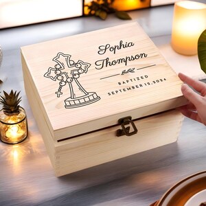 May include: A light-colored wooden box with a decorative cross and the name "Sophia Thompson" engraved on the lid. The box is secured with a metal clasp and the text "BAPTIZED SEPTEMBER 12, 2024" is also visible.