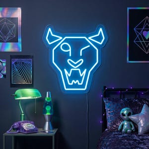 May include: A neon blue panther head light fixture winks on a dark blue wall. The room features geometric art prints, a green desk lamp, a purple rotary phone, and a lava lamp. A bed with a velvet comforter and decorative pillows is in the background.