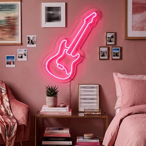 May include: A vibrant pink neon guitar outline on a pink wall, surrounded by framed pictures and small photographs. A small table with books and a plant sits beneath the sign, creating a stylish display.
