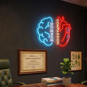 May include: A neon wall sign featuring a blue brain outline with the word SCIENCE and a red heart outline with the word COMPASSION. The sign is mounted on a dark wall above a desk with books, a small potted plant, and framed wall art.