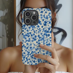 May include: A floral phone case featuring a pattern of small blue flowers on a cream background. The case has a camera cutout and is held in front of a person's face.