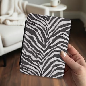 May include: A rectangular e-reader case featuring a high-contrast black and white zebra stripe pattern. The cover has a textured surface and a black spine edge, designed to protect the device while maintaining a consistent animal print aesthetic.