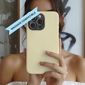 May include: A pale yellow phone case with a black camera module. The case is held up in front of a person's face. The text "MAGSAFE COMPATIBLE" is on a light blue banner across the top of the phone case.