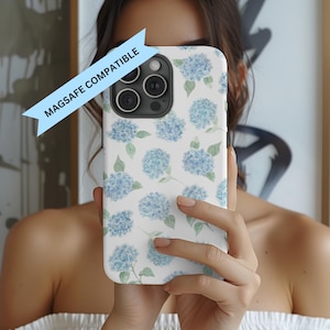 May include: White iPhone case with a blue hydrangea floral pattern. The case has a MagSafe Compatible label. The design features watercolor-style blue flowers and green leaves on a white background.