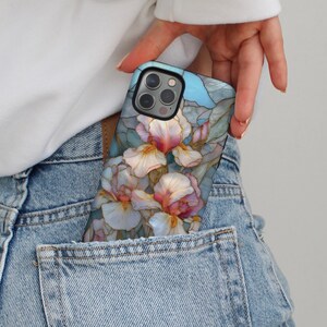 Könnte beinhalten: Eine Handyhülle mit floralem Design aus weißen und rosa Irisblüten und grünen Blättern auf hellblauem Hintergrund. Die Hülle befindet sich in einer Jeans-Tasche.