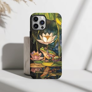 May include: A phone case with a stained glass design featuring a green frog sitting on a lily pad with water lilies and leaves. The case has a black border and a camera cutout.