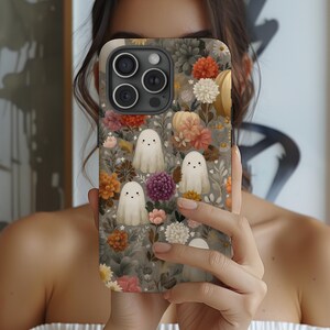 May include: A phone case featuring a floral pattern with white ghost illustrations. The case has a gray background with orange, pink, and white flowers. The phone case has a camera and flash.