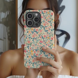 May include: A floral phone case featuring a cream background with a pattern of small pink, yellow, and blue flowers and green leaves. The case has a camera cutout and is held in front of a person.