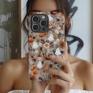 May include: A phone case featuring a floral pattern with orange, brown, and white flowers, pumpkins, and white ghost illustrations on a gray background. The case has a camera cutout and is held in front of a person.