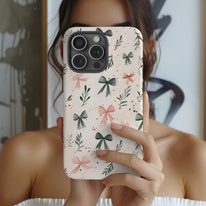 May include: A phone case with a cream background featuring a pattern of watercolor bows in pink and green, along with sprigs of leaves and small dots. The case has a camera cutout and is held up in front of a person.