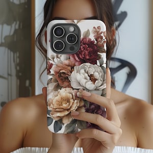 May include: A white phone case featuring a floral design with peonies and other flowers in shades of burgundy, peach, and cream. The case has a glossy finish and a camera cutout.