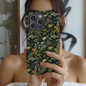 May include: A black smartphone case with a botanical pattern featuring green and yellow leaves and flowers. The case has a camera cutout and is held in front of a person's face.