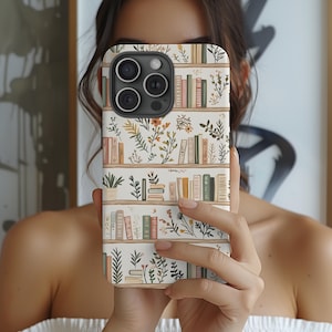 May include: A phone case featuring a bookshelf design with rows of books and floral accents. The case has a cream-colored background with a pattern of books in various colors, green plants, and orange flowers.