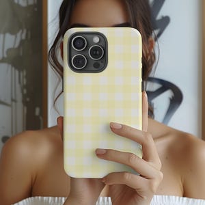 May include: A yellow and white gingham pattern phone case. The case is on a black phone with three camera lenses. The phone is held in front of a person's face.
