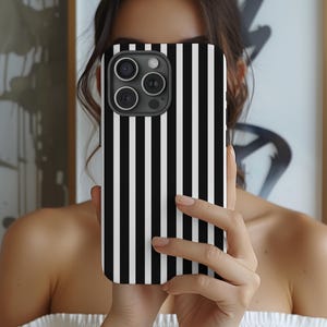 May include: A black and white striped phone case. The case has vertical stripes and a camera module with three lenses. The phone case is held in front of a person.