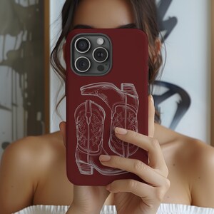 May include: A burgundy phone case featuring a white outline of two cowboy boots. The case has a camera lens cutout with three lenses.