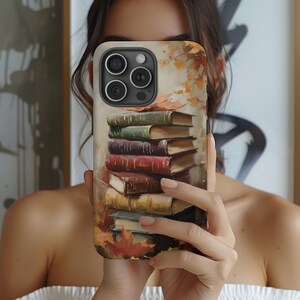 Könnte beinhalten: Eine Handyhülle mit einem Stapel Bücher im Herbst-Design. Die Hülle hat einen beigen Hintergrund mit bunten Herbstblättern und einem Stapel Bücher in verschiedenen Farben, darunter Grün, Rot und Braun.