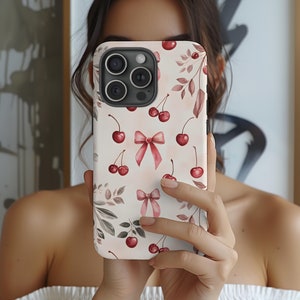 May include: A white phone case with a cherry and bow pattern. The case has a camera lens cutout and a pink bow design.