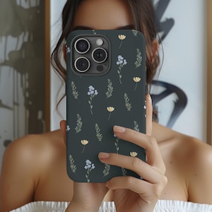 May include: A dark green phone case featuring a floral pattern with yellow and blue flowers and green leaves. The case has a camera cutout and is held up in front of a person's face.