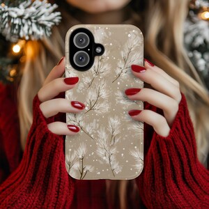 May include: A phone case with a beige background featuring a winter pine branch design. The case has a dual-lens camera cutout and is held by a person wearing a red sweater. The design includes white branches and small white dots.