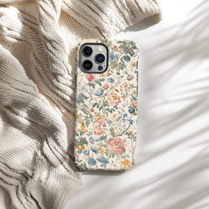 May include: A floral phone case featuring a cream background with a pattern of pink, blue, and yellow flowers and green leaves. The case has a black border and a camera cutout. The case is lying on a white surface next to a cream-colored knitted blanket.