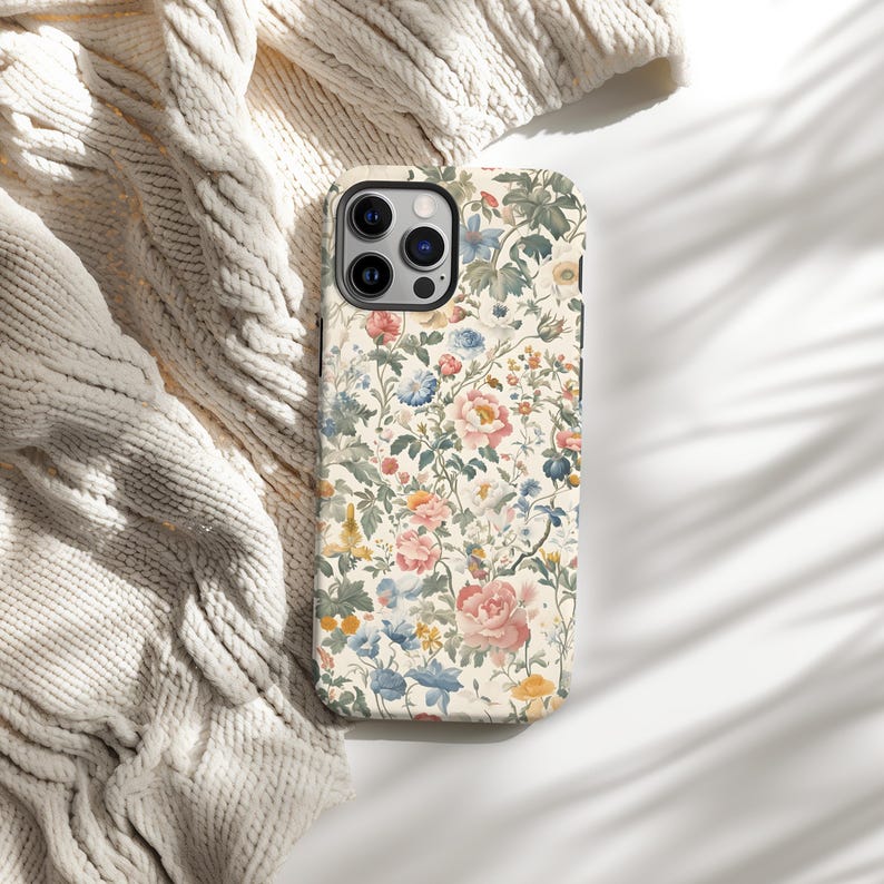 May include: A floral phone case featuring a cream background with a pattern of pink, blue, and yellow flowers and green leaves. The case has a black border and a camera cutout. The case is lying on a white surface next to a cream-colored knitted blanket.