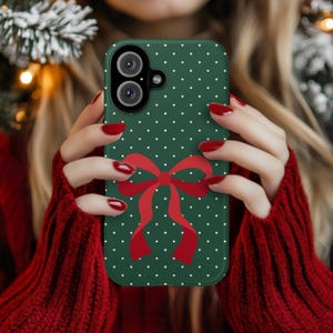 Peut inclure: Coque de téléphone verte à pois blancs et ornée d'un nœud rouge. Le boîtier présente une découpe pour un appareil photo à double objectif. L'arrière-plan est flou.