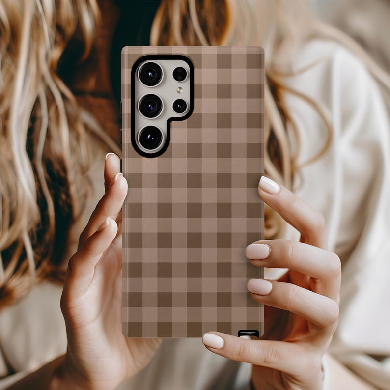 May include: A phone case with a brown and tan checkered pattern. The case has a camera cutout and a black button on the bottom. The phone case is being held by a person.