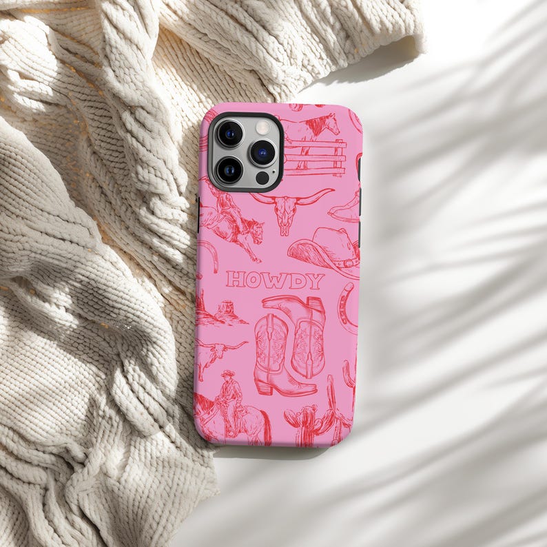 May include: Pink phone case with a repeating red cowboy-themed pattern. The design includes illustrations of horses, cowboy boots, hats, and the word "HOWDY". The case is shown on a cream-colored knitted surface.