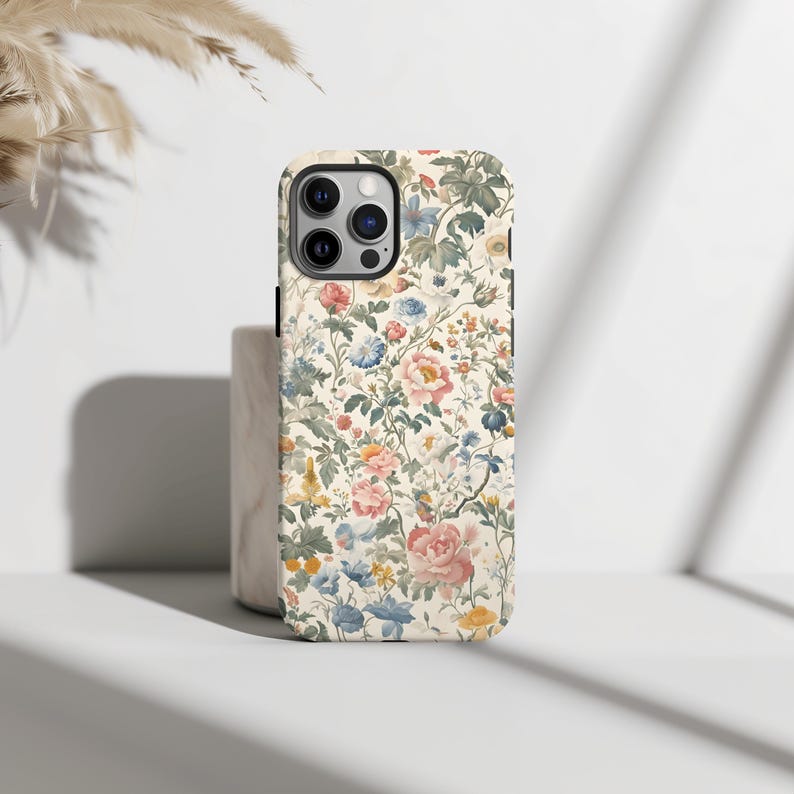 May include: A phone case with a floral pattern. The case features a cream-colored background with a repeating design of pink, blue, yellow, and green flowers and foliage. The case has a camera cutout and black edges.