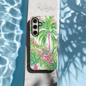 May include: A white phone case with a tropical print featuring pink flowers and green palm trees. The case has a cutout for a triple lens camera.